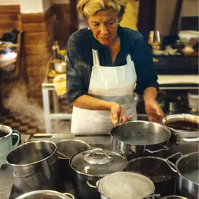 Christine Schauflinger mit weißer Schürze prüft dampfende Töpfe auf dem Herd.