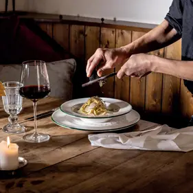 Servierperson reibt frische Trüffel über Pasta auf Teller, neben Rotweinglas, Kerze und Tischläufer auf rustikalem Holztisch.