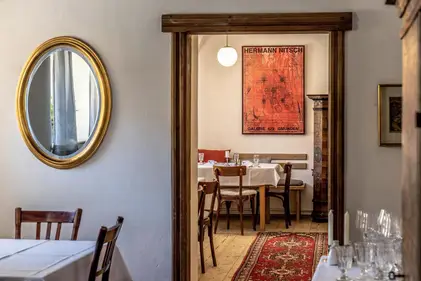 Gemütlicher Gastraum mit gedeckten Tischen, rotem Teppich und dem Poster HERMANN NITSCH an der Wand.