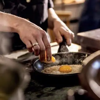 Hände brechen ein Ei über einer heißen Pfanne, ein Spiegelei brät und Dampf steigt auf.