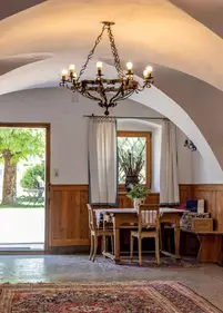 Rustikaler Essplatz unter Gewölbe mit hängendem Kronleuchter, Holztisch und Stühlen vor Fenster; offene Tür zum Garten.